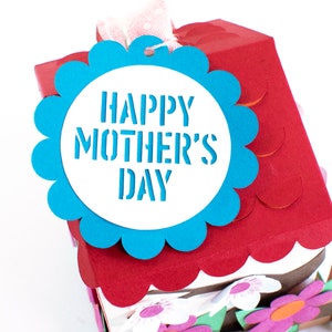 Op de afbeelding: Een blauw en wit papieren knutselhuis met een gegolfd dak en een blauw label met de tekst "Happy Mother's Day".