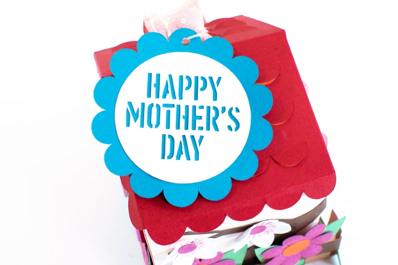 Op de afbeelding: Een blauw en wit papieren knutselhuis met een gegolfd dak en een blauw label met de tekst "Happy Mother's Day".