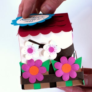 Op de afbeelding: Een rood en wit papieren vogelhuisje met een gegolfd dak en een blauw label met de tekst "Happy Mother's Day". Het vogelhuisje heeft twee witte bloemen met roze centra en een bruine tak. De basis van het vogelhuisje heeft twee roze bloemen met oranje centra en groene bladeren.