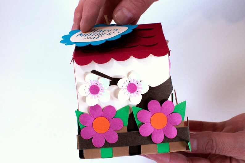 Op de afbeelding: Een rood en wit papieren vogelhuisje met een gegolfd dak en een blauw label met de tekst "Happy Mother's Day". Het vogelhuisje heeft twee witte bloemen met roze centra en een bruine tak. De basis van het vogelhuisje heeft twee roze bloemen met oranje centra en groene bladeren.