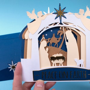 May include: A white paper Christmas card with a pop-up nativity scene. The scene features a stable with a manger, a shepherd, a camel, and a lamb. The card is decorated with gold stars and the words "Peace on Earth".