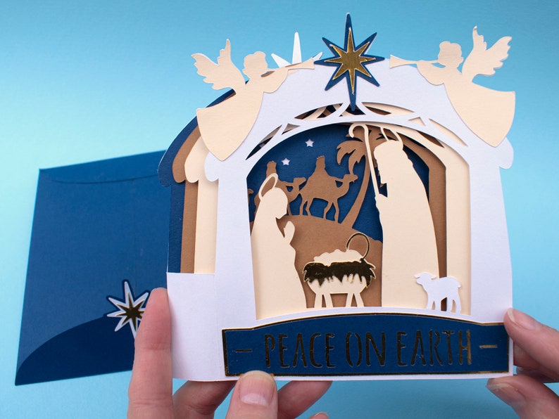 May include: A white paper Christmas card with a pop-up nativity scene. The scene features a stable with a manger, a shepherd, a camel, and a lamb. The card is decorated with gold stars and the words "Peace on Earth".