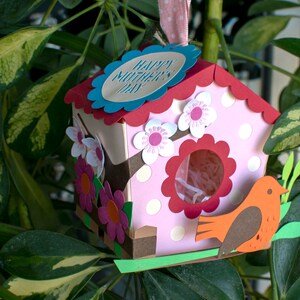 Op de afbeelding: Een roze en rood vogelhuisje met een gegolfd dak en een witte vogelhuisdeur. Het vogelhuisje heeft een blauw label met de tekst "Happy Mother's Day". Er zitten witte en roze bloemen op het vogelhuisje. Een kleine oranje vogel zit op een groene tak.