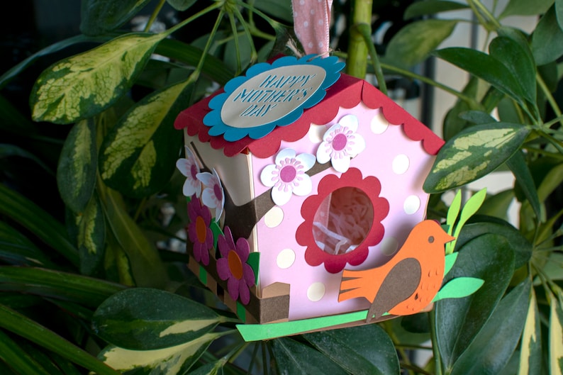 Op de afbeelding: Een roze en rood vogelhuisje met een gegolfd dak en een witte vogelhuisdeur. Het vogelhuisje heeft een blauw label met de tekst "Happy Mother's Day". Er zitten witte en roze bloemen op het vogelhuisje. Een kleine oranje vogel zit op een groene tak.