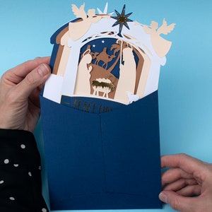 May include: A blue envelope with a pop-up Christmas card inside. The card features a nativity scene with a star, angels, and the text "Peace on Earth".