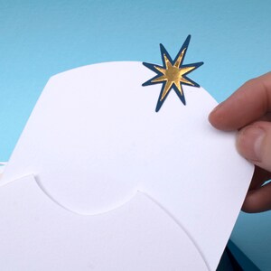 May include: A white card with a blue and gold star on the top right corner. The card is folded in half and has a scalloped edge.