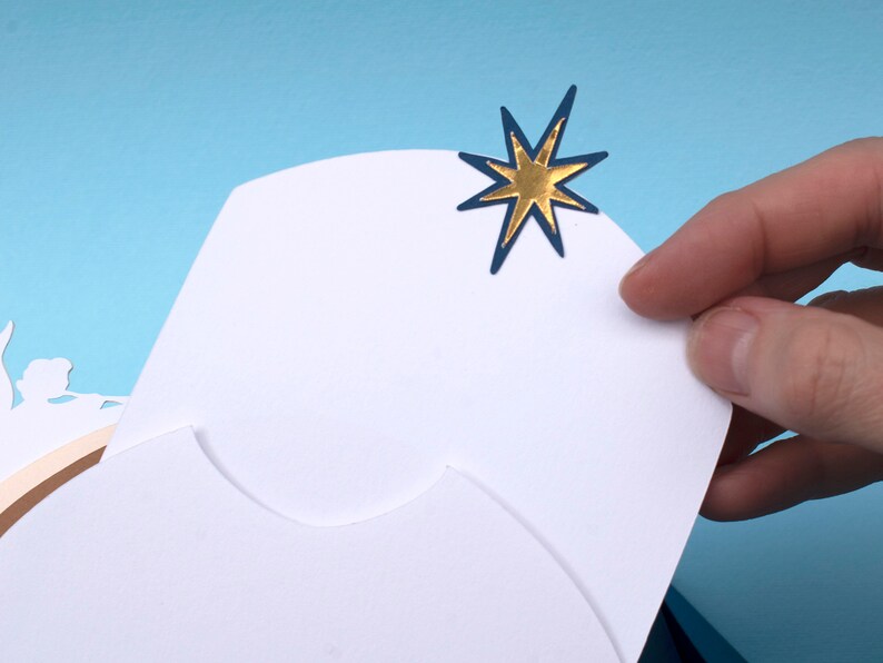 May include: A white card with a blue and gold star on the top right corner. The card is folded in half and has a scalloped edge.