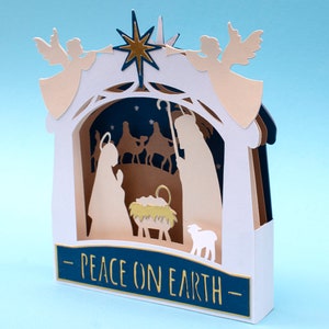 May include: A white papercraft nativity scene with a gold star, two angels, and the text "Peace on Earth".