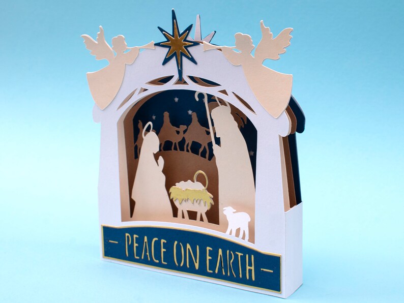 May include: A white papercraft nativity scene with a gold star, two angels, and the text "Peace on Earth".