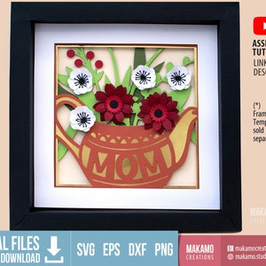 Op de afbeelding: Een rode en gouden theepot met het woord "MOM" aan de zijkant, omringd door witte en rode papieren bloemen. De theepot bevindt zich in een zwart frame met een witte passe-partout.