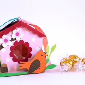 Op de afbeelding: Een roze en rood vogelhuisje met witte stippen en een bruine vogel die op de rand zit. Het vogelhuisje heeft een gegolfd dak en een kleine opening waar de vogel naar binnen kan. Op de voorgrond liggen drie gouden chocolade-eieren.