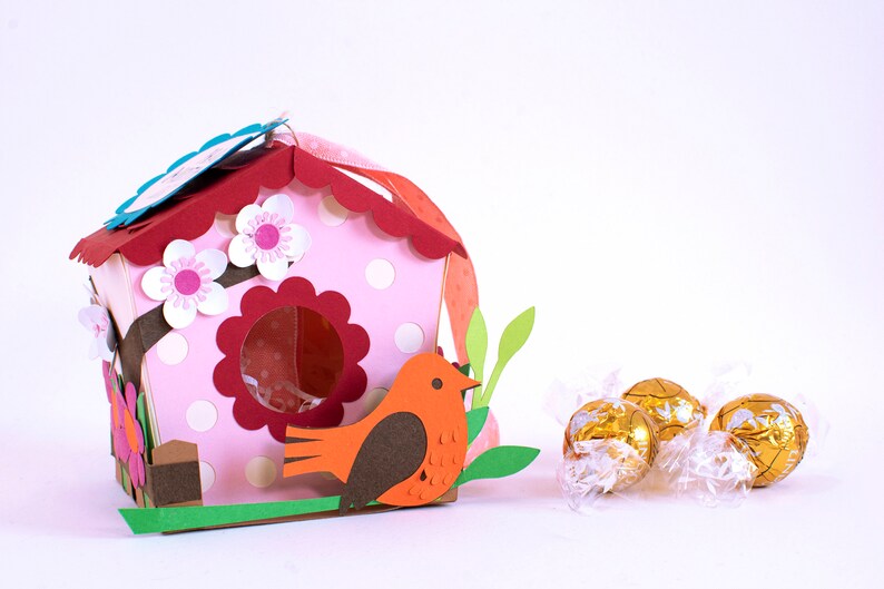 Op de afbeelding: Een roze en rood vogelhuisje met witte stippen en een bruine vogel die op de rand zit. Het vogelhuisje heeft een gegolfd dak en een kleine opening waar de vogel naar binnen kan. Op de voorgrond liggen drie gouden chocolade-eieren.