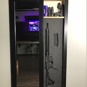 May include: A black gun safe with a mirrored door, revealing a rifle and other firearms inside. The safe is mounted on a wall and has a black frame.