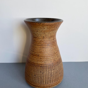 May include: A handcrafted, speckled brown ceramic vase with a dark brown interior. The vase features a flared top and a textured, ribbed design around the body, with vertical lines near the base. A decorative piece for home decor.