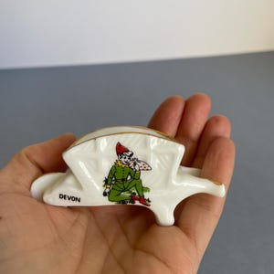 May include: A white ceramic souvenir ashtray with gold detailing. The ashtray features a cartoon figure in green and red clothing, holding a bag. The word "DEVON" is printed on the side. A decorative item.