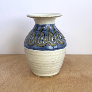 May include: A ceramic vase with a cream base and a blue band featuring gold swirl designs. The vase has a wide, textured body and a narrow neck. The rim is cream-coloured.