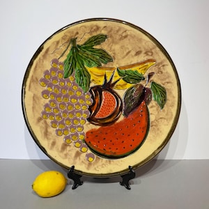 May include: A round ceramic plate with a painted fruit design. The plate features a bunch of grapes, a slice of watermelon, a pomegranate, and bananas. The plate is on a black stand, and a lemon is next to the stand.