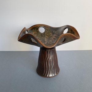 May include: A brown ceramic decorative bowl with a pedestal base. The bowl has a wavy rim with circular cutouts and a textured, tree-trunk-like base. The interior of the bowl has a green and brown pattern.