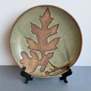 May include: A round ceramic plate with a leaf design. The plate has a light green background with a large brown leaf imprint in the centre. The rim of the plate is a darker brown. The plate is displayed on a black stand.