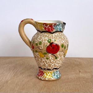 May include: A small, hand-painted ceramic jug with a tan base, decorated with a red rose and colourful floral accents. The handle is tan with red dots. White squiggly lines and black trim add detail to the design.