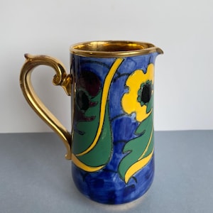 May include: A blue ceramic jug with a gold handle and rim. The jug is decorated with yellow flowers, green leaves, and black accents. The design is set against a blue background.