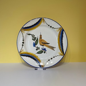 May include: A white ceramic plate with a blue and yellow border. The centre of the plate features a hand-painted yellow bird with blue accents perched on a green branch with blue flowers.