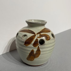 May include: A small, white ceramic vase with brown floral designs and holes around the top. The vase has a rounded shape and a slightly flared lip.