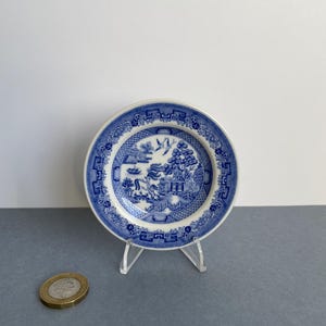May include: A small, round blue and white plate featuring a Willow pattern design. The plate is displayed on a clear plastic stand. A coin is next to the plate. The plate has a detailed landscape scene with a bridge, trees, and buildings.