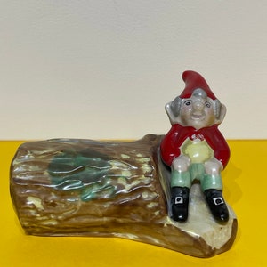 May include: A ceramic figurine of a gnome wearing a red hat and sitting on a brown tree stump. The gnome is wearing green trousers and black shoes.