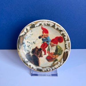 May include: A round decorative plate depicting a winter scene with a bear and two gnomes. One gnome plays an instrument, the other holds a bowl. The plate has a white border with a gold and brown design.