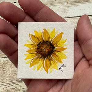 May include: A small square watercolor painting of a single sunflower with vibrant yellow petals and a dark brown center. The artwork features a textured white paper background and includes the artist's signature in the bottom right corner.