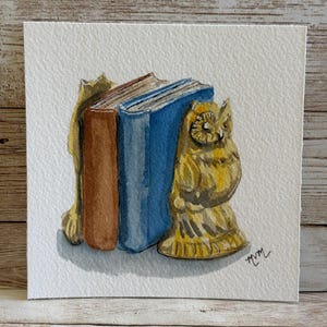 May include: Watercolor painting of a stack of books held by a gold cat bookend and a gold owl bookend. The books are brown and blue. The artwork is on white paper.