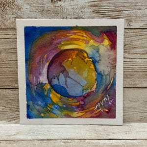 May include: Watercolor painting on a square white paper. The artwork features a central circular shape with a gradient of colors, including yellow, purple, blue, and orange. The artist's signature is in the lower right corner.