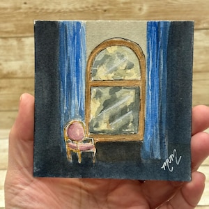 May include: Watercolor painting of a window with a wooden frame, blue curtains, and a small pink chair. The artwork is on a square piece of paper and held in a hand. The painting has a signature in the bottom right corner.