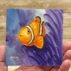 May include: Watercolor painting of an orange clownfish with white stripes, set against a blue and purple background. The fish is the focal point, with the artist's signature in the lower left corner. The artwork is on a square piece of paper.