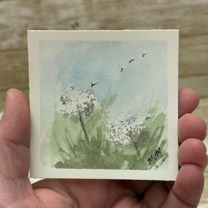 May include: A small watercolor painting of dandelions in a field. The artwork features white dandelion seed heads, green grass, and a light blue sky with birds. The artist's signature "Mury" is in the bottom right corner.