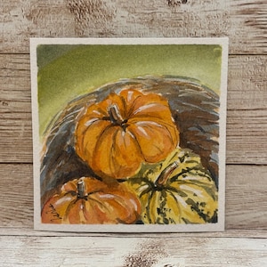 May include: A watercolor painting on paper depicting three pumpkins. One large orange pumpkin sits centrally, with a smaller orange pumpkin to the left and a yellow-green speckled pumpkin to the right. The background is a soft green wash.