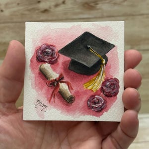 May include: Watercolor painting of a graduation theme. A black mortarboard with a gold tassel, a rolled diploma tied with a red ribbon, and three burgundy roses are on a square canvas with a pink background.