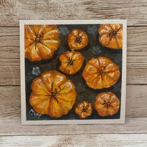May include: A square watercolor painting on white paper featuring seven orange pumpkins of varying sizes arranged on a dark, muted green background. Each pumpkin has detailed black ink stems and vines, with the artist's signature MVM in the corner.