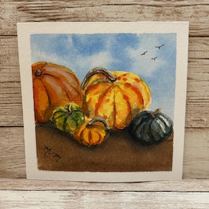 May include: A watercolor painting on square paper depicting a collection of pumpkins in various sizes and colors, including orange, green, and dark gray, set against a light blue sky with three small black birds flying in the distance.