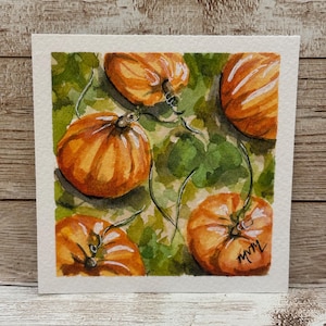 May include: A square watercolor painting on textured white paper featuring four orange pumpkins nestled among green foliage. The pumpkins are depicted with visible stems and vines, and the artist's signature is written in black ink in the bottom right.