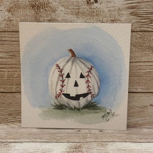 May include: A watercolor painting on square paper depicting a white pumpkin carved with a jack-o'-lantern face. The pumpkin features red baseball-style stitching patterns on its sides, set against a soft blue circular wash background.