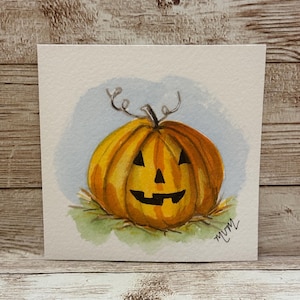 May include: A square watercolor painting on textured paper featuring a bright orange jack-o'-lantern with a carved black face, a green stem, and curly tendrils. The pumpkin sits on a patch of green grass against a light blue background.