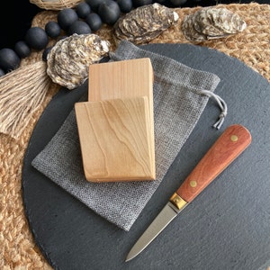May include: An oyster knife with a wooden handle and brass accents, on a dark grey slate board. Two light-coloured wooden blocks and a grey drawstring bag are also present. The knife blade has the word "SPEENSON" etched on it.