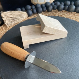 May include: An oyster knife with a wooden handle and a stainless steel blade lies on a dark grey surface. A light-coloured wooden oyster shucking box is open in the background. The knife is designed for safely opening oysters.