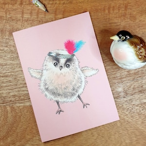 Puede incluir: Una tarjeta rosa con un dibujo de un pequeño pájaro blanco y esponjoso que lleva un sombrero con plumas rosas y azules. El pájaro tiene ojos grandes y oscuros y está de pie sobre dos patas.