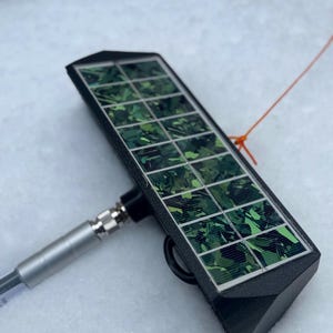 May include: A small, rectangular solar panel with a black frame and a grid of green and black cells. A silver cable and connector are attached to the panel. An orange string is tied to the panel.