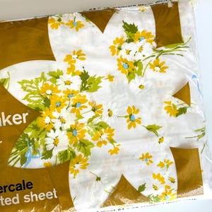 May include: A packaged percale fitted sheet with a floral pattern. The sheet features a white background with yellow, white, and blue daisy-like flowers and green leaves. The packaging is a clear plastic bag with a gold-colored border.