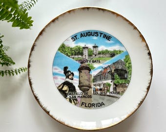 May include: A white decorative plate with a gold rim featuring a colorful illustration of St. Augustine, Florida. The plate includes images of the City Gate, Castillo de San Marcos, and the Oldest House.