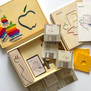 May include: A vintage Macintosh computer software kit in a yellow box, including several 3.5-inch floppy disks, a spiral-bound manual, stickers with the rainbow Apple logo, and printed documentation featuring line art of the original computer.